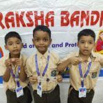 Three schoolboys in beige uniforms wear medals and id lanyards posing in front of a raksha bandhan banner